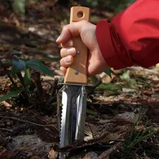 Nisaku NJP650 hori hori Japanese weeding knife being pushed into leaf litter by a gloveless hand in a red sleeve, the wooden handle and the concave serrated stainless blade both visible