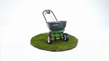 Scotts Elite Spreader photographed on a white studio background, standing on a circular disc of turf, its twin-disc spreading mechanism visible beneath the hopper