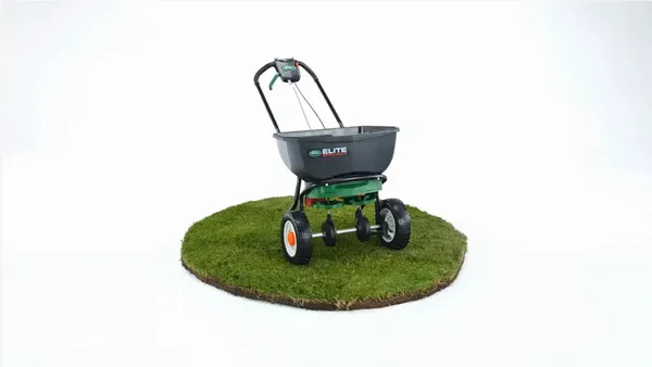 Scotts Elite Spreader photographed on a white studio background, standing on a circular disc of turf, its twin-disc spreading mechanism visible beneath the hopper