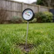 Long-probe analogue soil moisture meter pushed into lawn soil with its dial facing the camera — XLUX moisture meter review